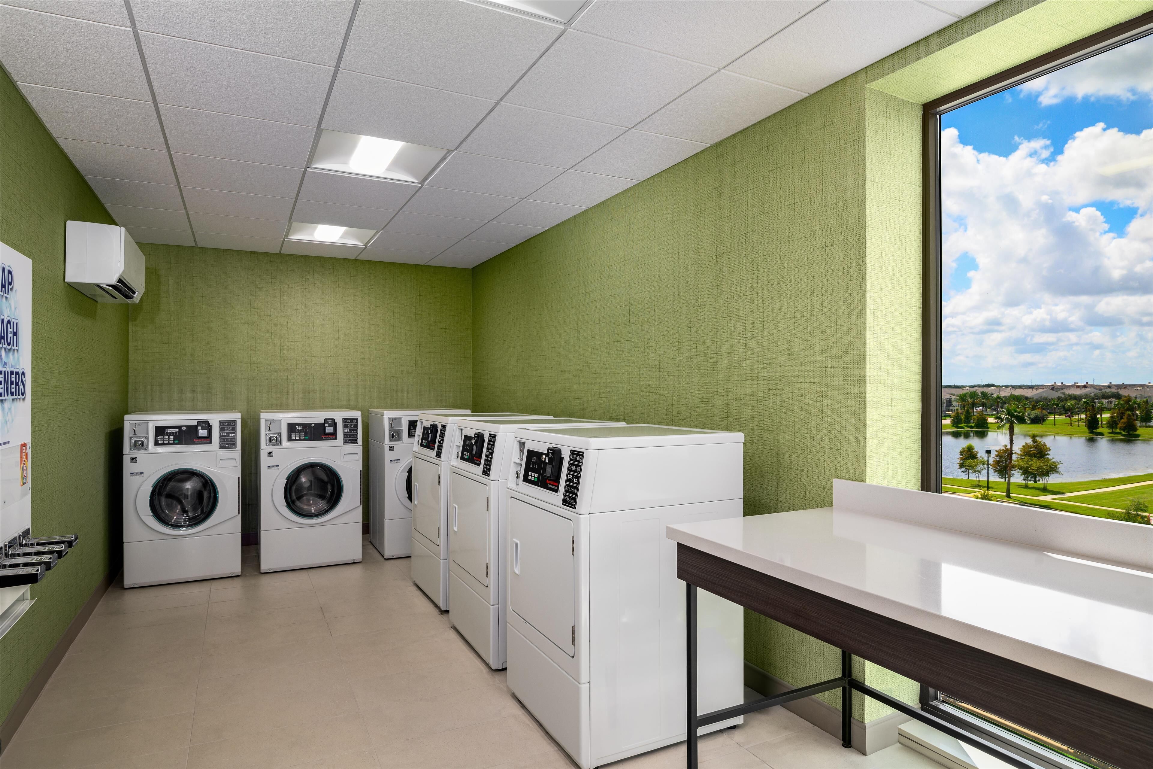 A laundry room with four white washing machines, a large window with a view of a park, and a white counter.