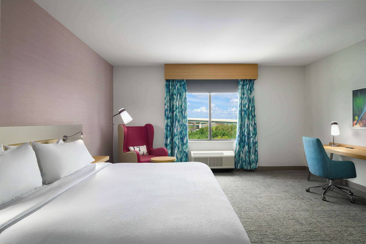 Ein Hotelzimmer mit Bett, Schreibtisch, Stuhl, einem Fenster mit Vorhängen und einem Bild an der Wand.