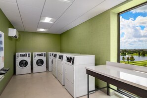 A laundry room with four front-loading washing machines, a drying rack, and a large window offering a view of a lake and greenery. - Hilton Garden Inn Millenia near Universal Orlando Resort (Orlando)