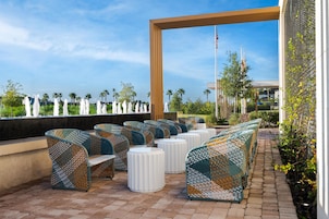 Outdoor seating with woven chairs and white stools, a clear sky, and a view of palm trees.