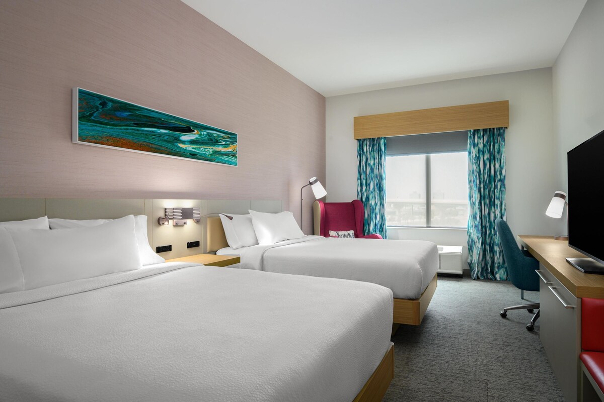 A hotel room with two beds, a desk, a TV, a window with curtains, and a painting on the wall.
