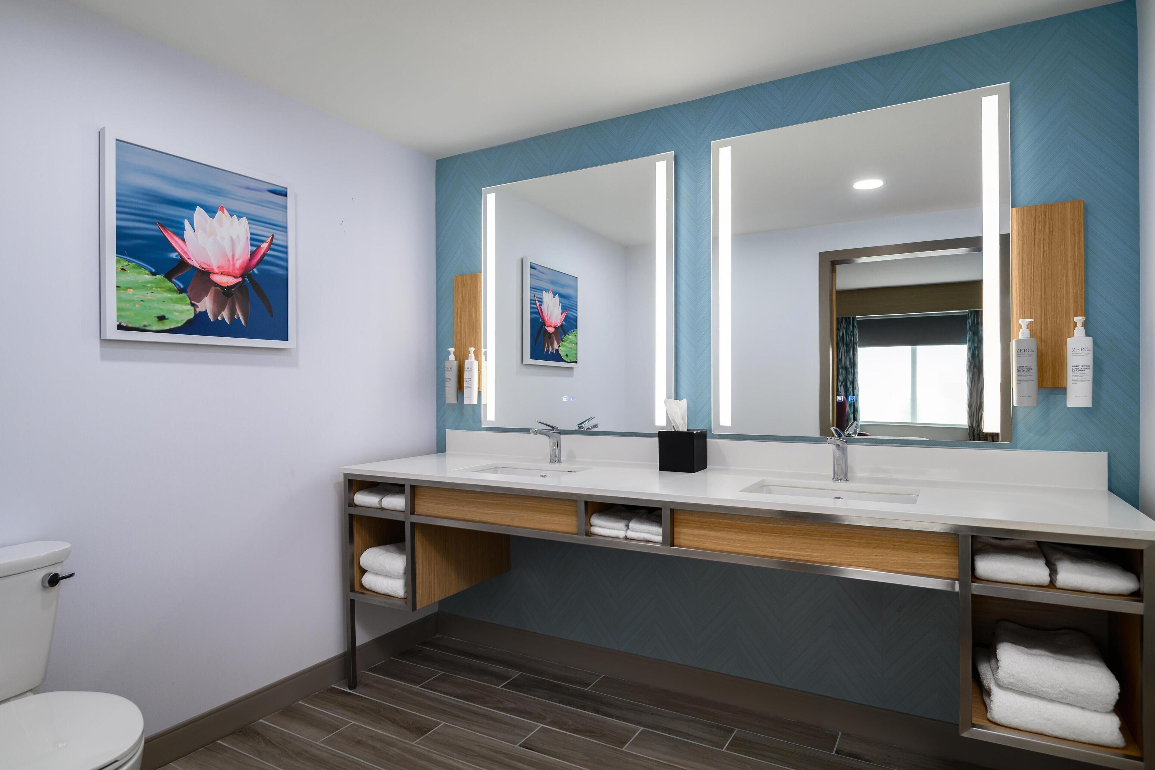A modern bathroom with a large mirror, two sinks, and a toilet. There are towels and toiletries on the counter.