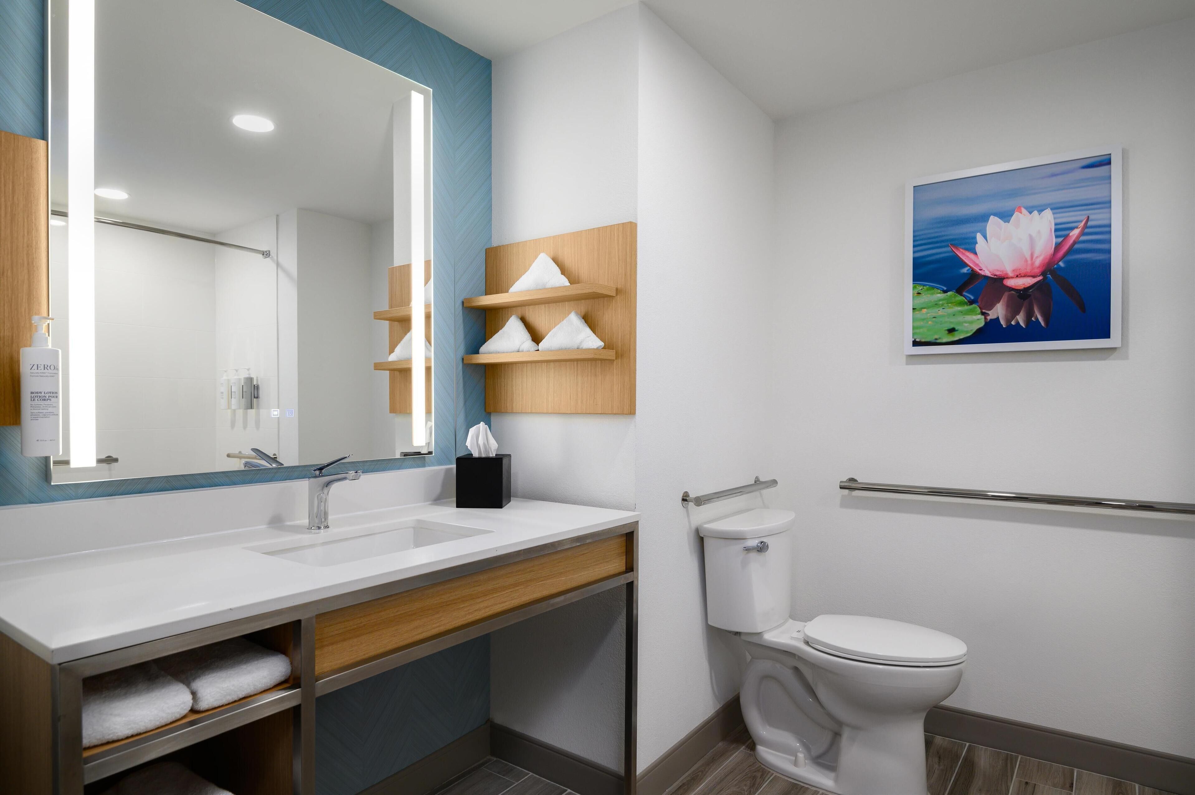 Een moderne badkamer met een grote spiegel, een witte wastafel en toilet. Daarboven hangen houten planken bij de wastafel en aan de muur een schilderij van een lotusbloem.