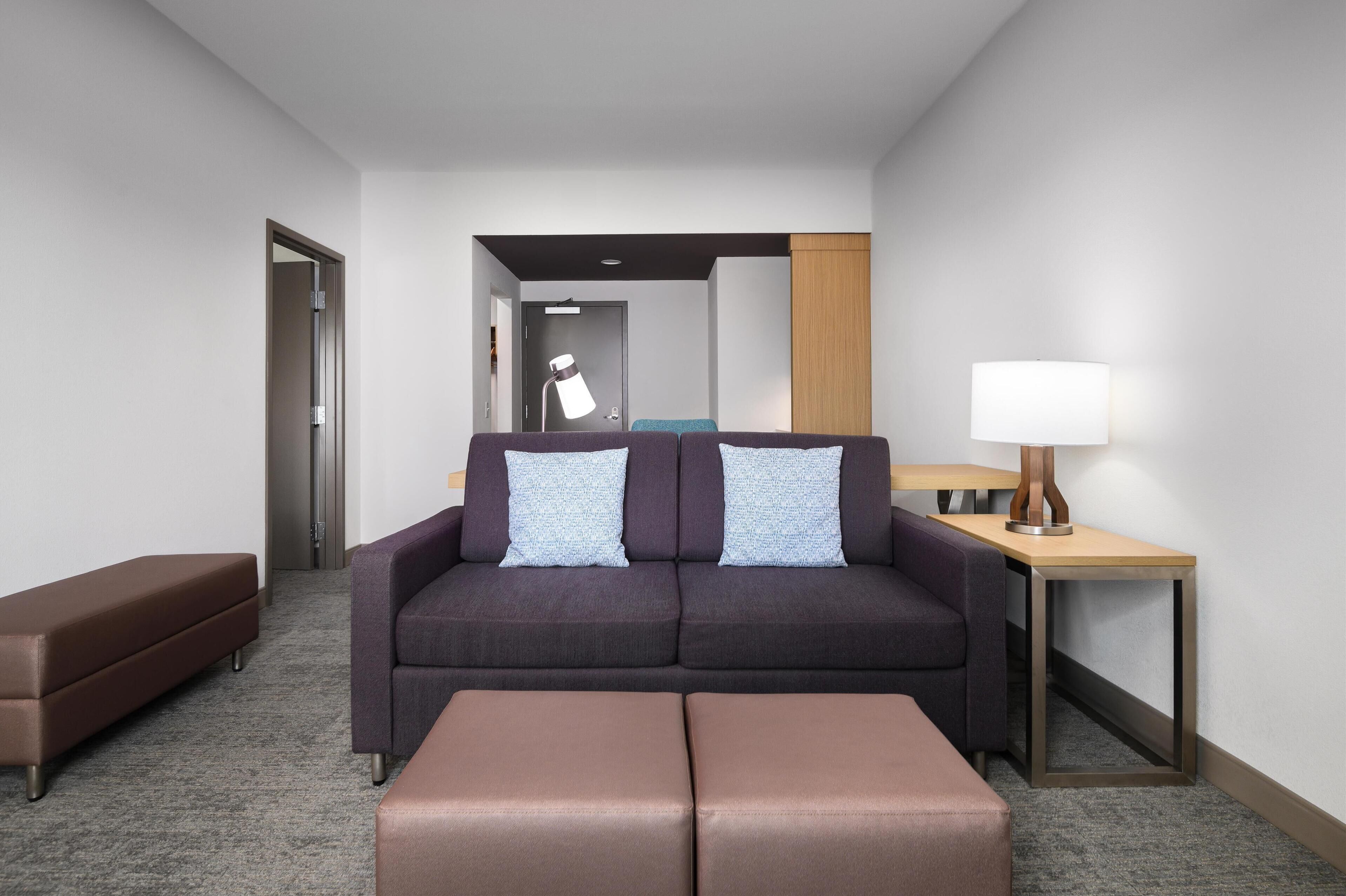 Quarto de hotel moderno com sofá, duas almofadas, mesa lateral com luminária e um banco.
