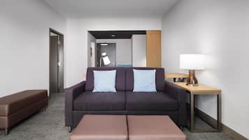 Quarto de hotel moderno com sofá, duas almofadas, mesa lateral com luminária e um banco.