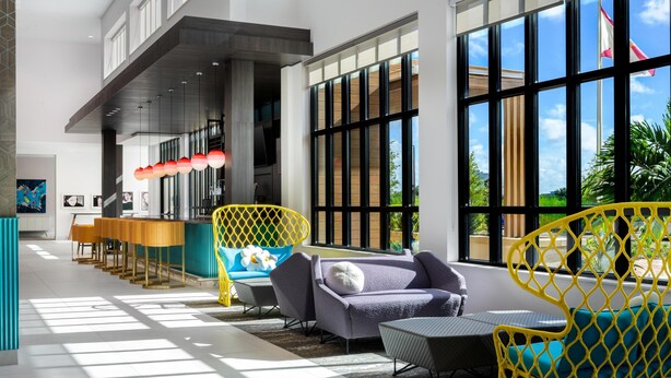 Un hall d'hôtel moderne avec un comptoir d'accueil, un espace salon doté de chaises jaunes et bleues, et de grandes fenêtres offrant une vue sur l'extérieur.