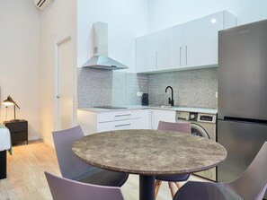 Une cuisine compacte avec des meubles blancs, un réfrigérateur en acier inoxydable, un four encastré et un évier. On y trouve une table ronde avec des chaises et une petite table d’appoint avec une lampe.