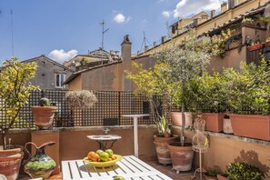 A rooftop terrace with potted plants, a table with a bowl of fruit, and a birdcage.