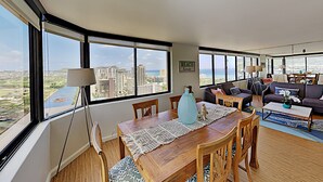 Ein geräumiges Wohnzimmer mit einem großen Esstisch, bequemen Sitzgelegenheiten und Blick auf Stadt und Meer durch große Fenster.