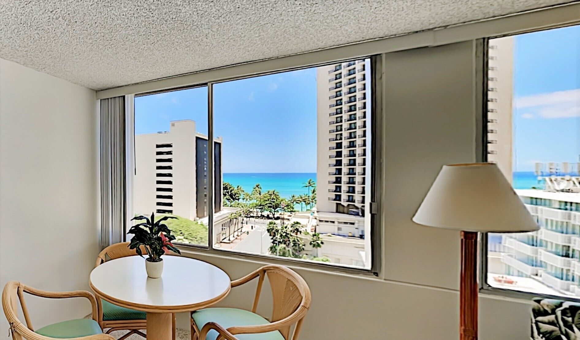 A hotel room with a large window offering a view of the ocean and tall buildings. There is a round table with chairs and a potted plant.