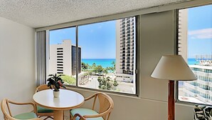 A hotel room with a large window offering a view of the ocean and tall buildings. There is a round table with chairs and a potted plant. - Pacific Monarch Paradise 1-Bedroom | Ocean Views (Honolulu)