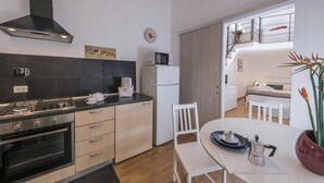 Et moderne køkken med gasblus, ovn og mikroovn. Der er et spiseområde med et hvidt bord og stole, samt udsigt til et soveværelse med seng og et natbord.