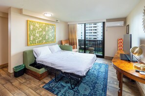 A hotel room with a bed, a desk, a chair, a small table, a trash bin, and a window with a view of buildings.