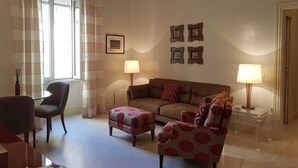 A living room with a sofa, two armchairs, a coffee table, and a side table with a lamp. There are curtains on the window, and a framed picture on the wall. - Deluxe flat 2 bedrooms (Roma)