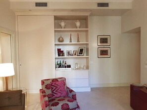 Uma sala de estar com sofá, mesa lateral, luminária, estante com objetos decorativos e quadros nas paredes.