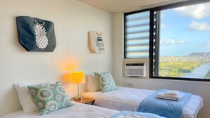 Quarto de hotel com duas camas, janela com vista para a cidade, ar condicionado fixado na parede e uma luminária.