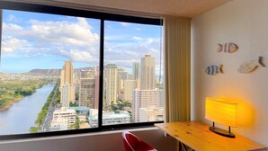 Quarto de hotel com uma janela grande que oferece vista para a cidade, uma escrivaninha de madeira com luminária, uma cadeira vermelha e arte de parede com tema de peixes decorativos.