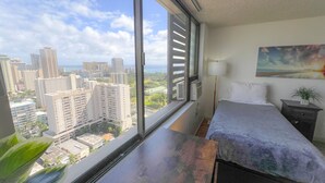 A bedroom with a large window, a bed, a bedside table, a lamp, and a painting on the wall. - Bright Hawaiian Monarch Studio | Full Kitchen (Honolulu)