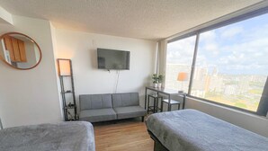 A hotel room with two beds, a sofa, a TV, a desk with a chair, a lamp, and a view of the city. - Bright Hawaiian Monarch Studio | Full Kitchen (Honolulu)