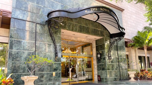 L’entrée d’un hôtel, avec une façade en marbre, une enseigne indiquant « The Royal Garden », et une porte en verre donnant sur l’intérieur.