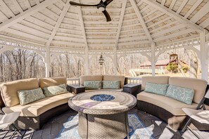 Relax in a cozy outdoor space with a fire pit, sofas, and armchairs, surrounded by a pergola and lush greenery.