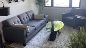 Ett modernt vardagsrum med en grå soffa, ett soffbord i glas och ett stort fönster som släpper in naturligt ljus.
