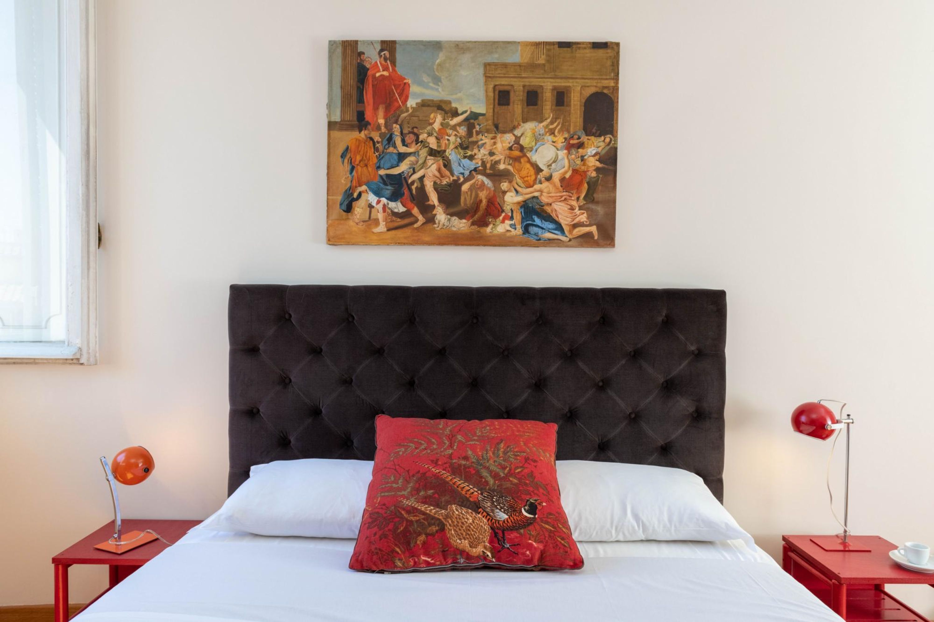 A bedroom with a tufted headboard, a red pillow with a bird design, a painting of a historical scene, and a red lamp on a nightstand.