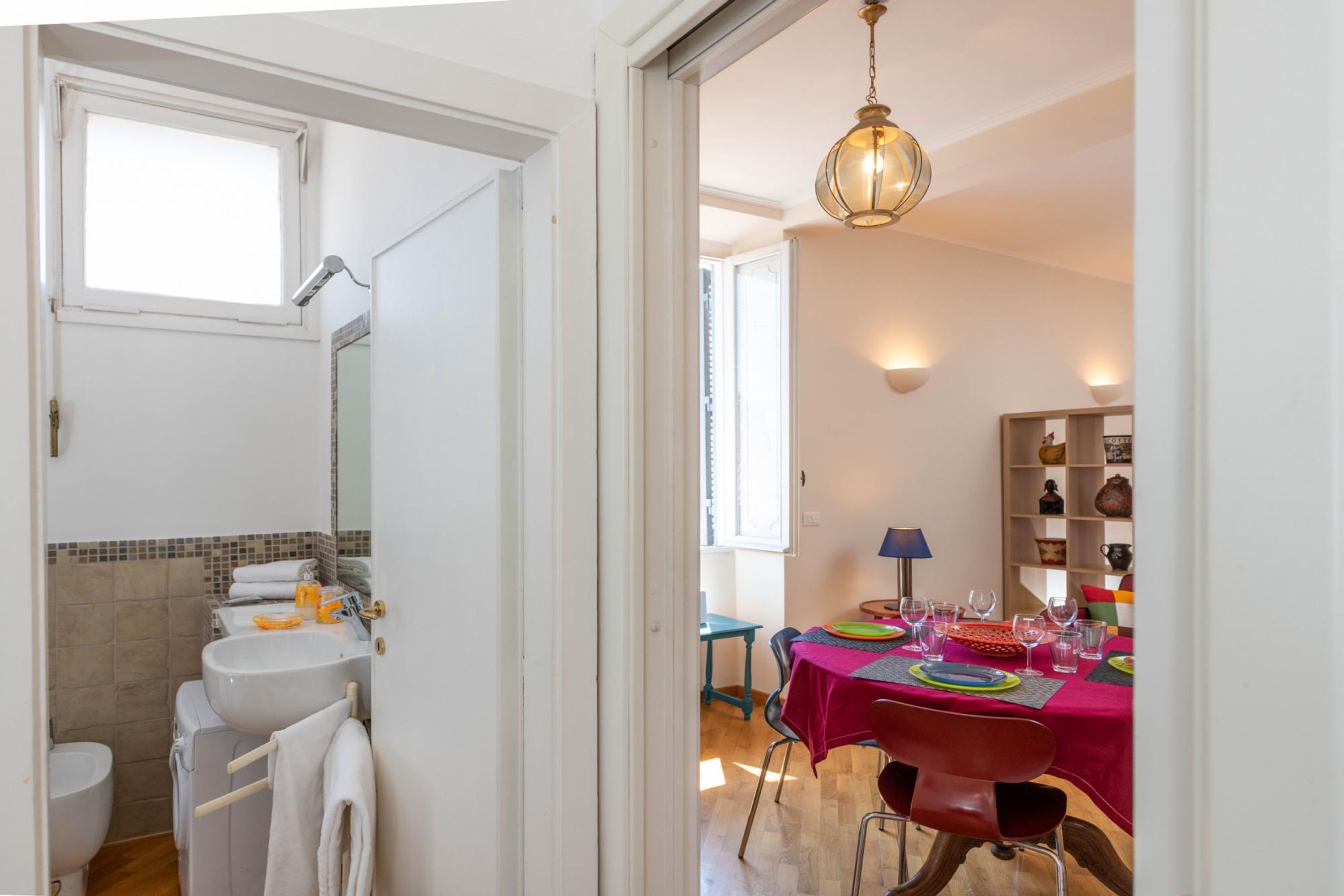 A bathroom with a sink, mirror, and towel rack. There is a dining area with a table set for four, chairs, and a chandelier.