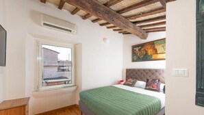 Een slaapkamer met een houten plafond, een raam met uitzicht op gebouwen, een bed met een groene deken en een schilderij aan de muur.