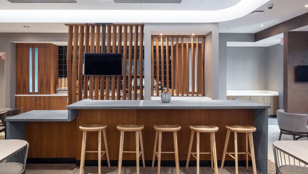 A modern bar area with a wooden bar counter, four stools, a flat-screen TV mounted on the wall, and a large window providing natural light. - SpringHill Suites Tucson at The Bridges (Tucson)