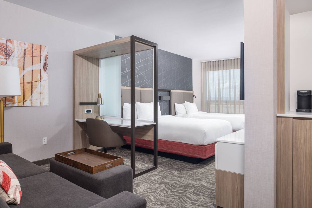 A modern hotel room with a bed, a desk, a chair, a sofa, and a coffee machine.
