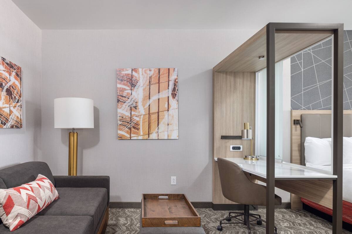 A hotel room with a grey sofa, a desk, a chair, a bed, and two artworks on the wall.