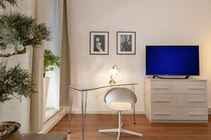 A modern living room with a glass desk, a white chair, a television, and two framed pictures on the wall.
