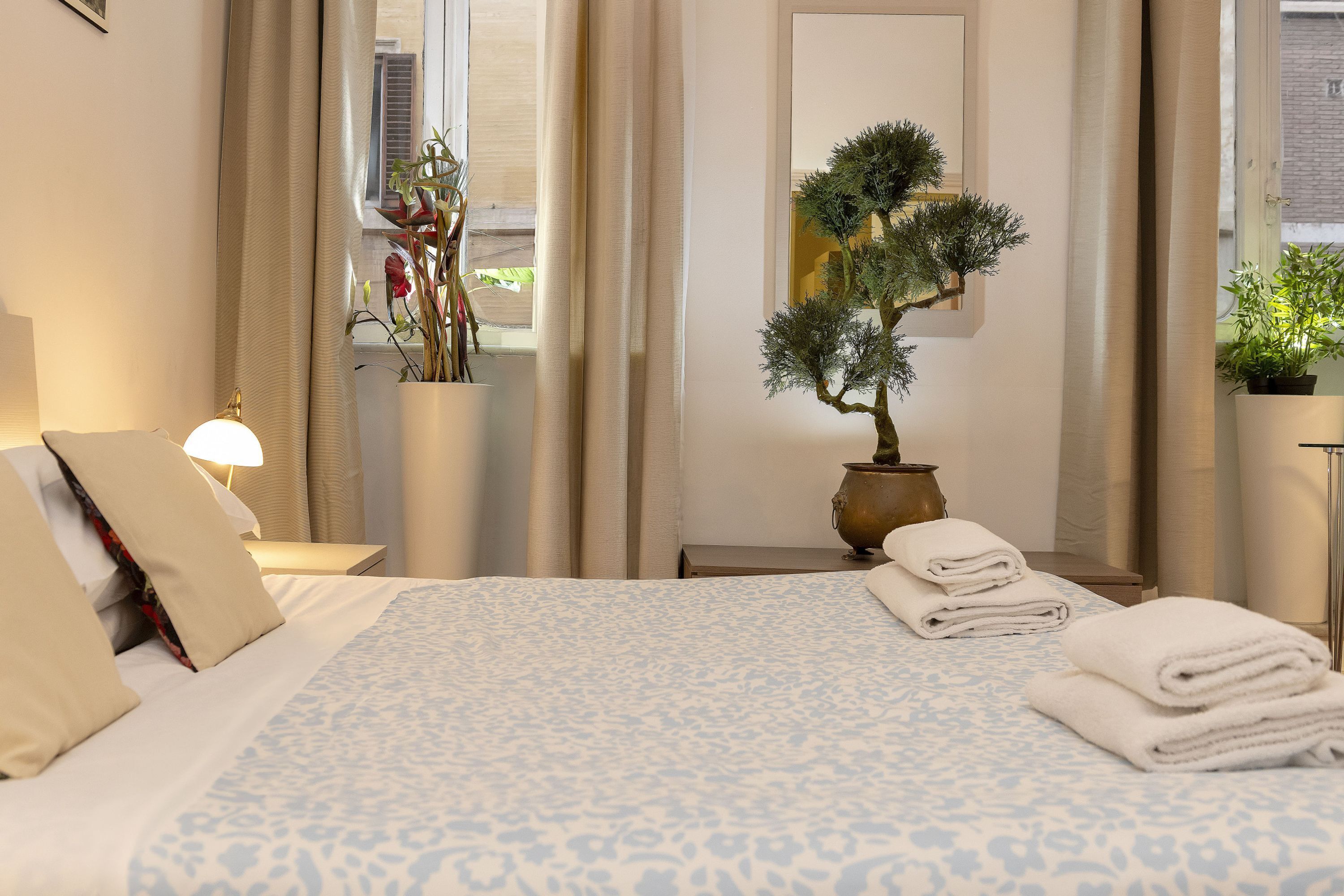 A hotel room with a bed, a nightstand, a lamp, a potted plant, and a window with curtains.