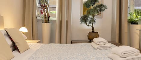 Habitación de hotel con cama, mesita de noche, lámpara, planta en maceta y ventana con cortinas.