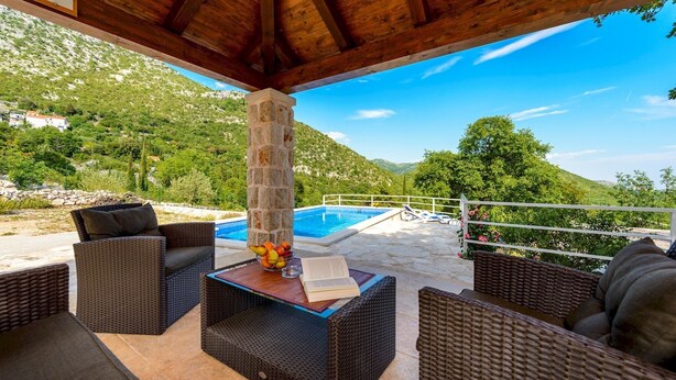 Un espace extérieur aménagé avec des meubles en osier, un pilier en pierre et une vue sur une piscine et des montagnes.