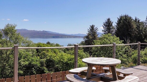 A wooden deck with a picnic table and benches overlooking a lake with mountains in the distance. - The Lookout (Sleat)