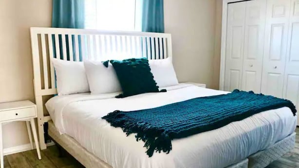 A neatly made bed with white bedding and a teal throw blanket, a white nightstand with a drawer, and a white wardrobe in a room with wooden flooring.