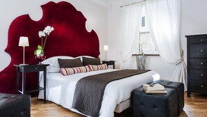 A modern bedroom with a large bed, a red accent wall, a black nightstand, a white lamp, and a window with white curtains. - Corso Vittorio - Campo de Fiori (Rome)