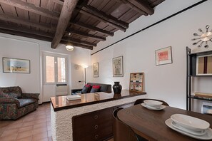 A cozy living room with a sofa, a coffee table, and a dining table with plates and bowls. There are framed pictures on the walls and a clock above the dining table. - Cedro House in Trastevere (Rome)
