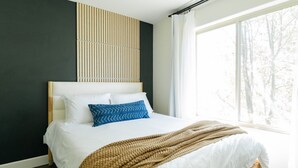 A neatly made bed with white linens and a blue patterned pillow, a brown knitted throw, and a wooden headboard.
