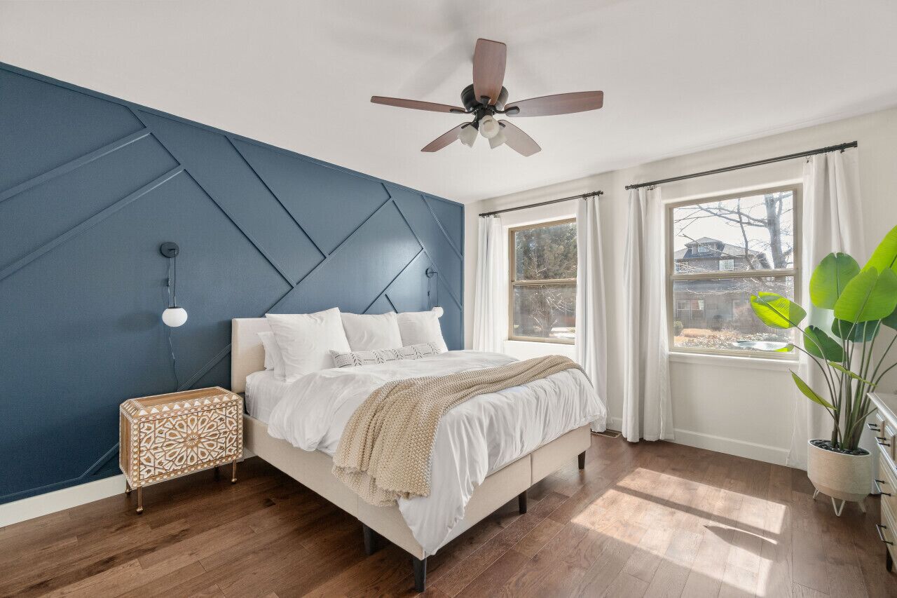 A bedroom with a bed, a ceiling fan, a nightstand, a window with curtains, and a potted plant.