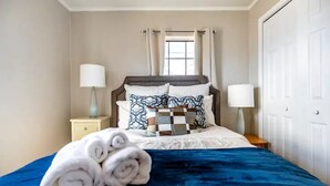 Un letto rifatto con cura, una coperta blu e asciugamani arrotolati, una lampada da comodino e una finestra con tende.