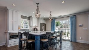 Une cuisine moderne avec des meubles blancs, un îlot central doté de tabourets de bar, un éclairage suspendu et une grande fenêtre ornée de rideaux bleus.