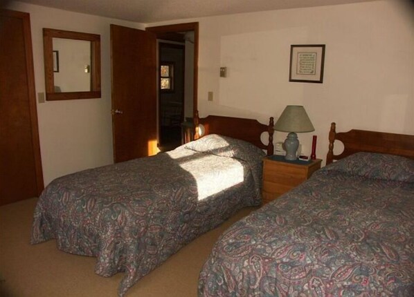 A room with two beds, a nightstand with a lamp, a framed picture on the wall, and a door leading to another room.
