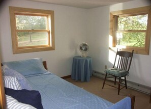 A bedroom with a bed, a chair, a table with a fan, and two windows with wooden frames.