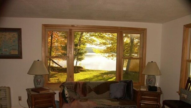 A cozy living room with a large window offering a view of a lake surrounded by trees. There is a wooden sofa with a blanket and pillows, two wooden side tables with lamps, and a framed picture on the wall. - Wendell Cottage (Belgrade)