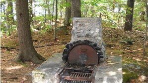 A rustic stone fireplace with a metal grate, set in a forested area with trees and a stone base. - Haskell's Lodge on Long Pond! (Belgrade)