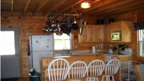 A wooden cabin kitchen with a refrigerator, stove, and hanging pots. There are four white chairs around a small dining table. - Sandy Beach Cottage! (Belgrade)