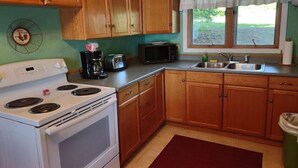 A kitchen with wooden cabinets, a white stove, a microwave, a coffee maker, and a window with a view of greenery. - Gramp's Camp (Belgrade)
